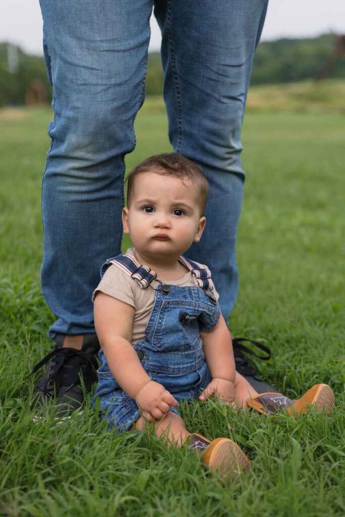A baby wearing blue jean overalls, looking into the camera sitting near his fathers feet on a grassy field