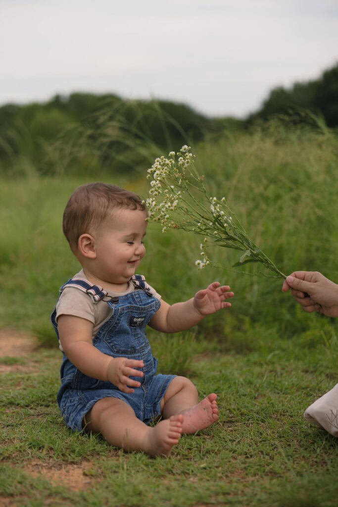 A baby sitting next to his mom holding some wild flowers smiling during a family photography session