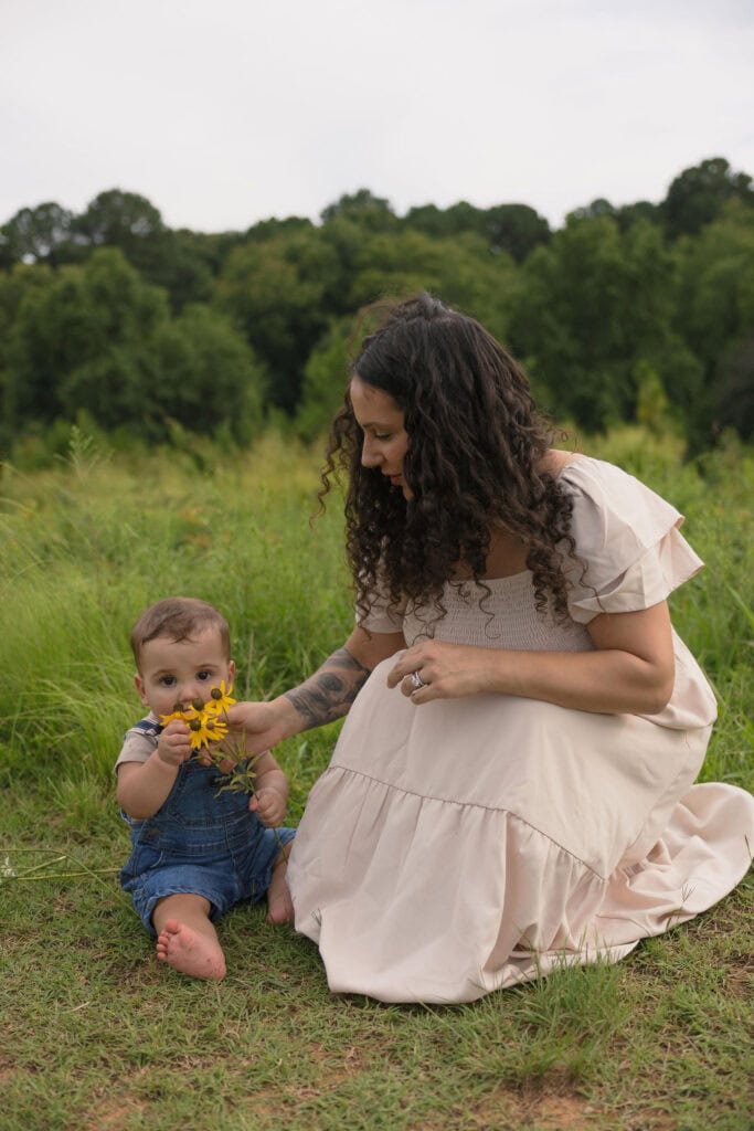 A baby sitting next to his mom while holding yellow wild flowers amongst a field of wild flowers