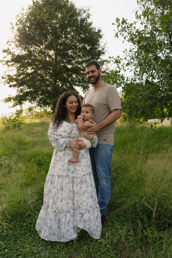 A posed photograph of a family holding their 9 month of baby in front of some trees at golden hour standing amongst tall grass