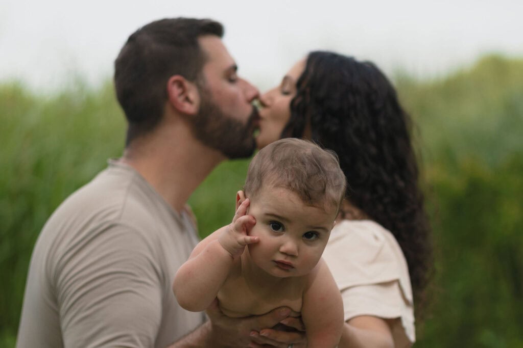 Parents kissing in the background which they are holding up their baby that is looking into the camera