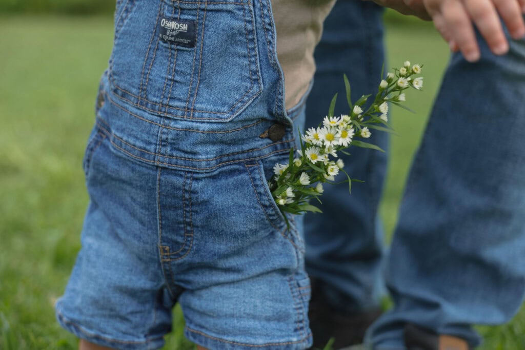 A close up photograph of a toddler wearing blue jean overalls with wild flowers tucked into his pocket
