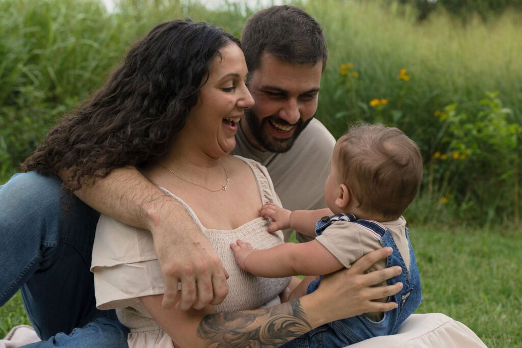 Parents holding their 9 month old baby while smiling and looking at them. They are standing in front of some tall green grass at the NC Museum of Art