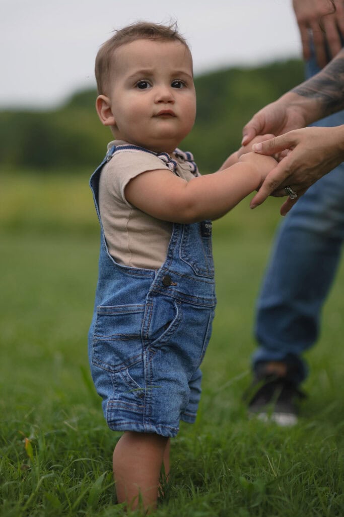 Baby wearing jean overalls looking at the camera while holding his moms hands in a family photography session at the NCMA in Raleigh, NC