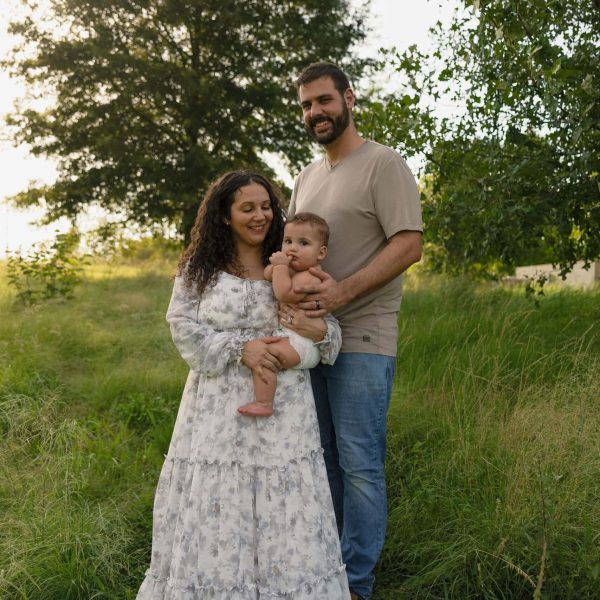 A posed photograph of a family holding their 9 month of baby in front of some trees at golden hour standing amongst tall grass