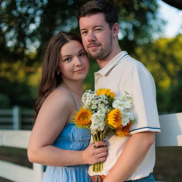 Couple looking into the camera posed for their couples session with Amelia and Matt Photo at Historic Oak View Park. They are leaning on a white picket fence leaning on each other holding flowers