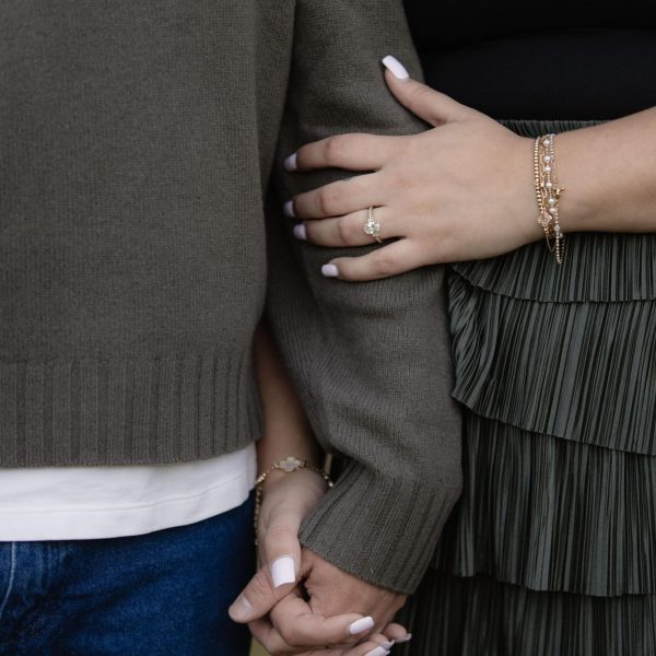a couple holding eachother's hands while standing near eachother. The woman's engagement ring is showing and she is wearing several bracelets. They are posing for their engagement photos and this was a detail shot taken by Amelia and Matt photo